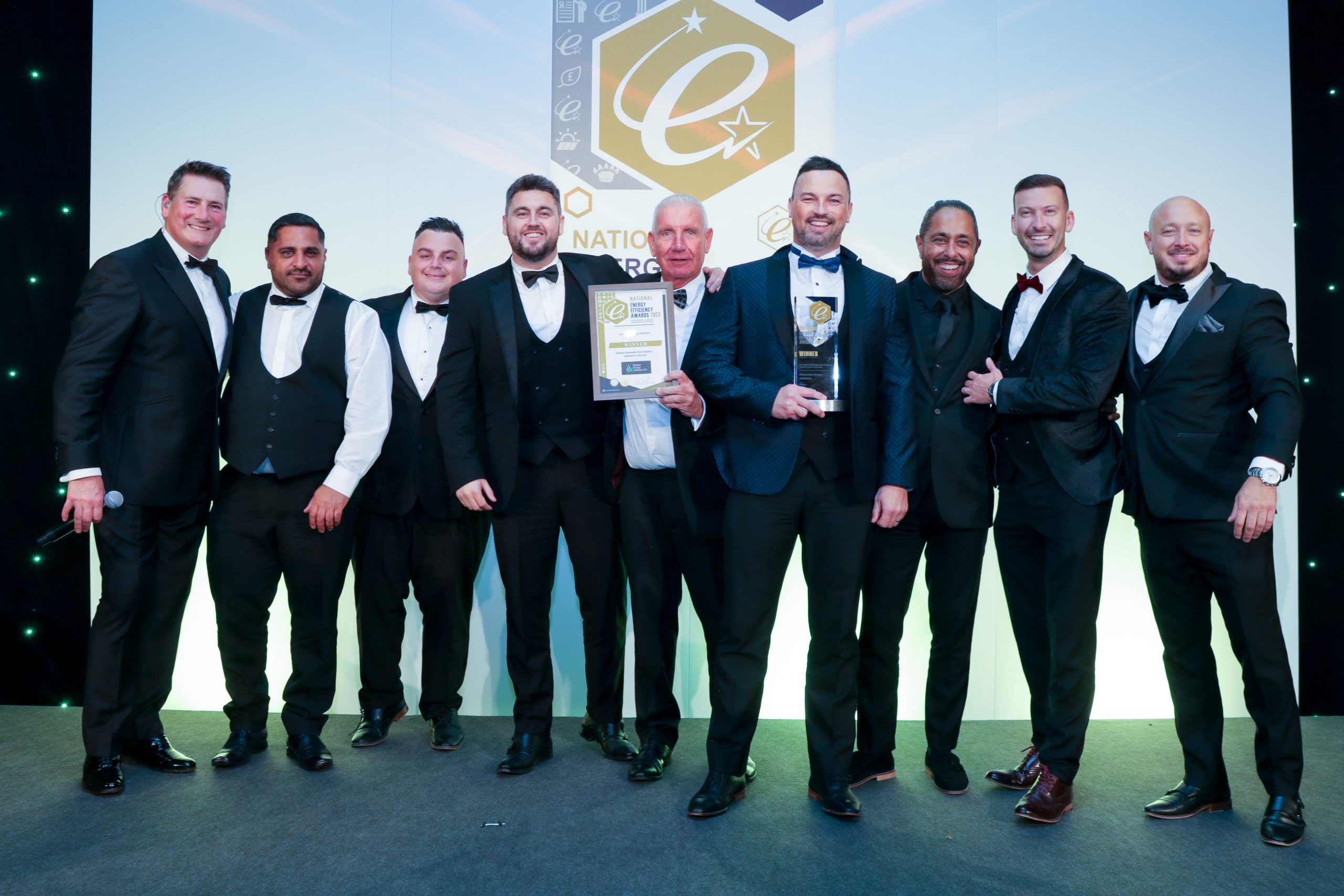 A group of men in tuxedos lined up on stage smiling whilst holding two awards.