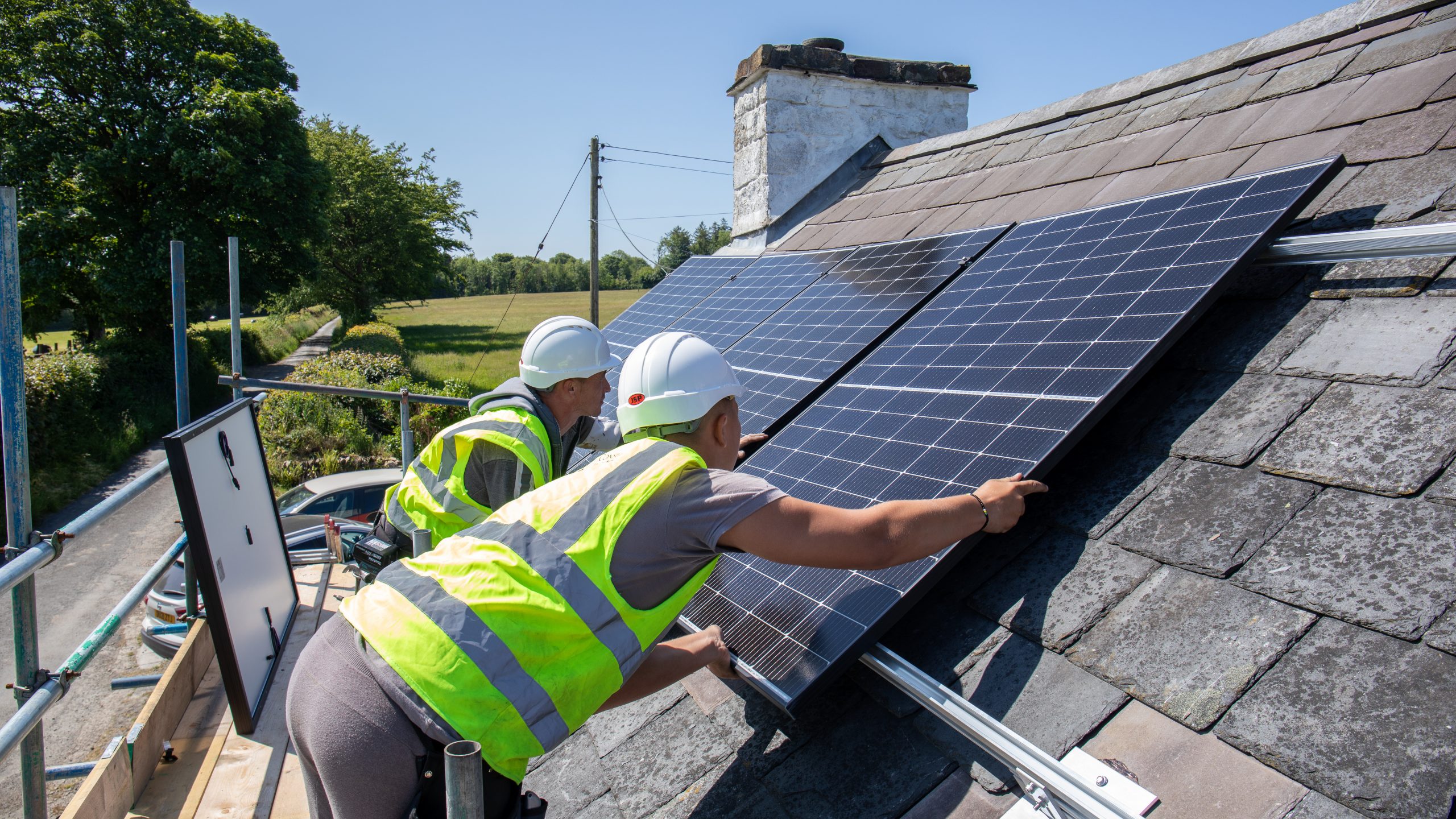 Two workers in high-vis vests and helmets fitting solar panels on the roof of a house.