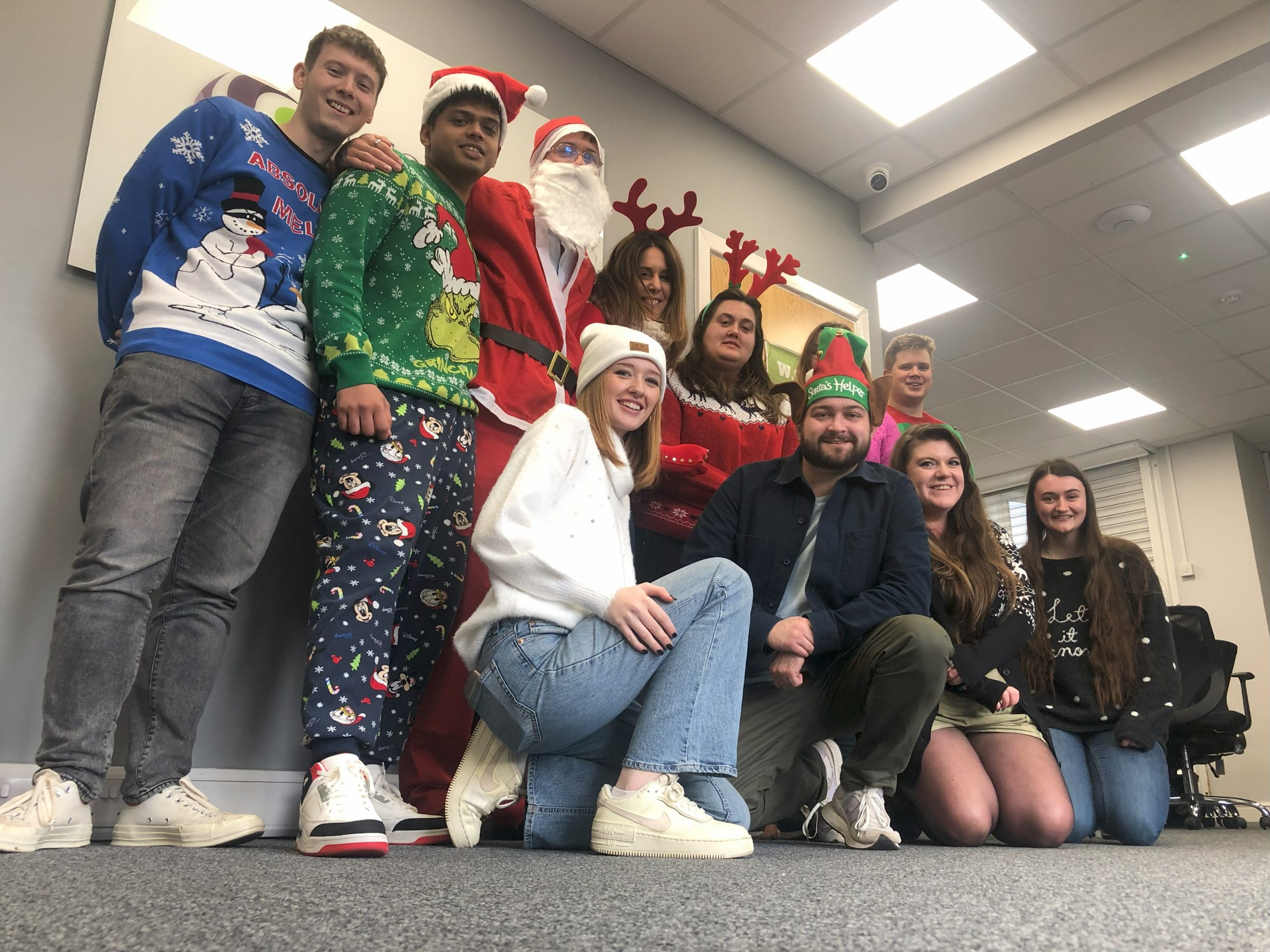 A group of people standing together smiling wearing Christmas jumpers and accessories.